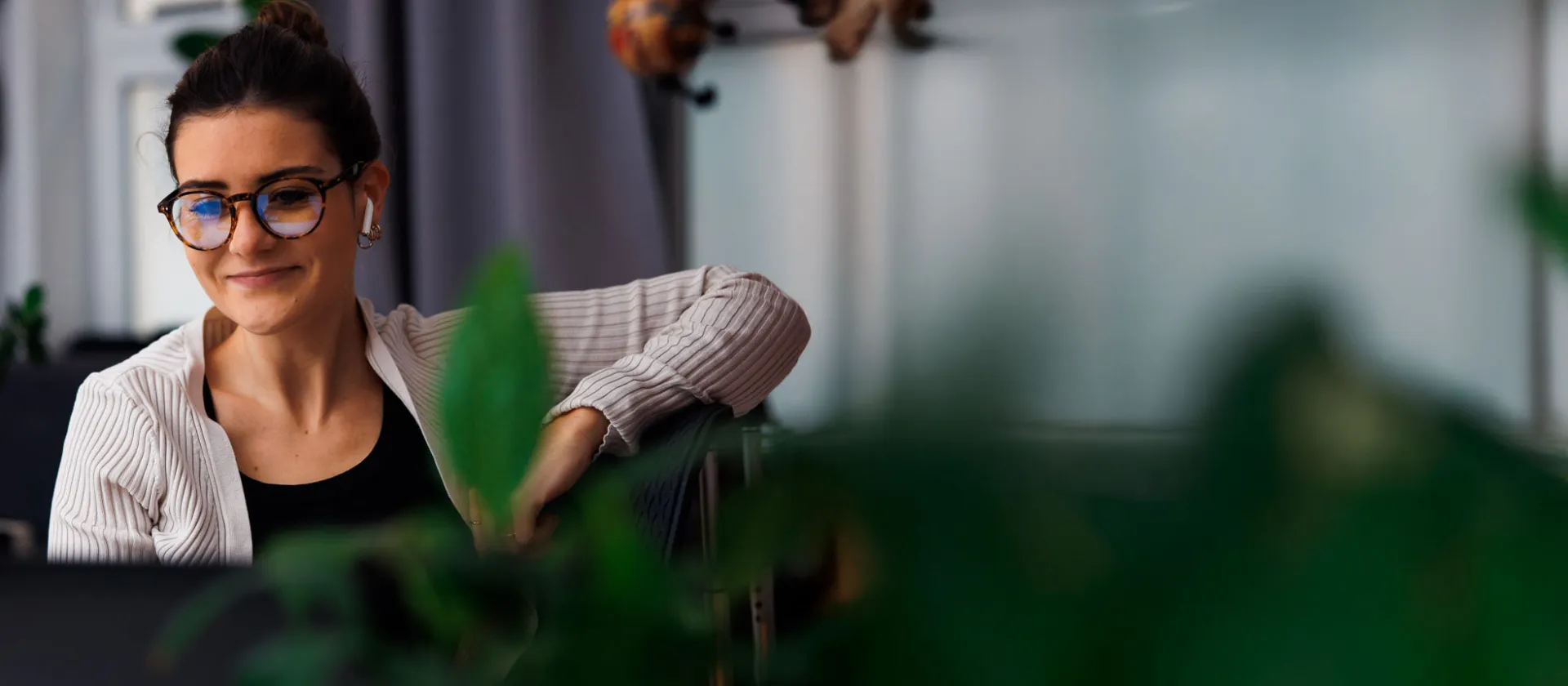 woman with glasses working remotely, smiling while engaged in a virtual meeting, surrounded by greenery in a modern office setting.