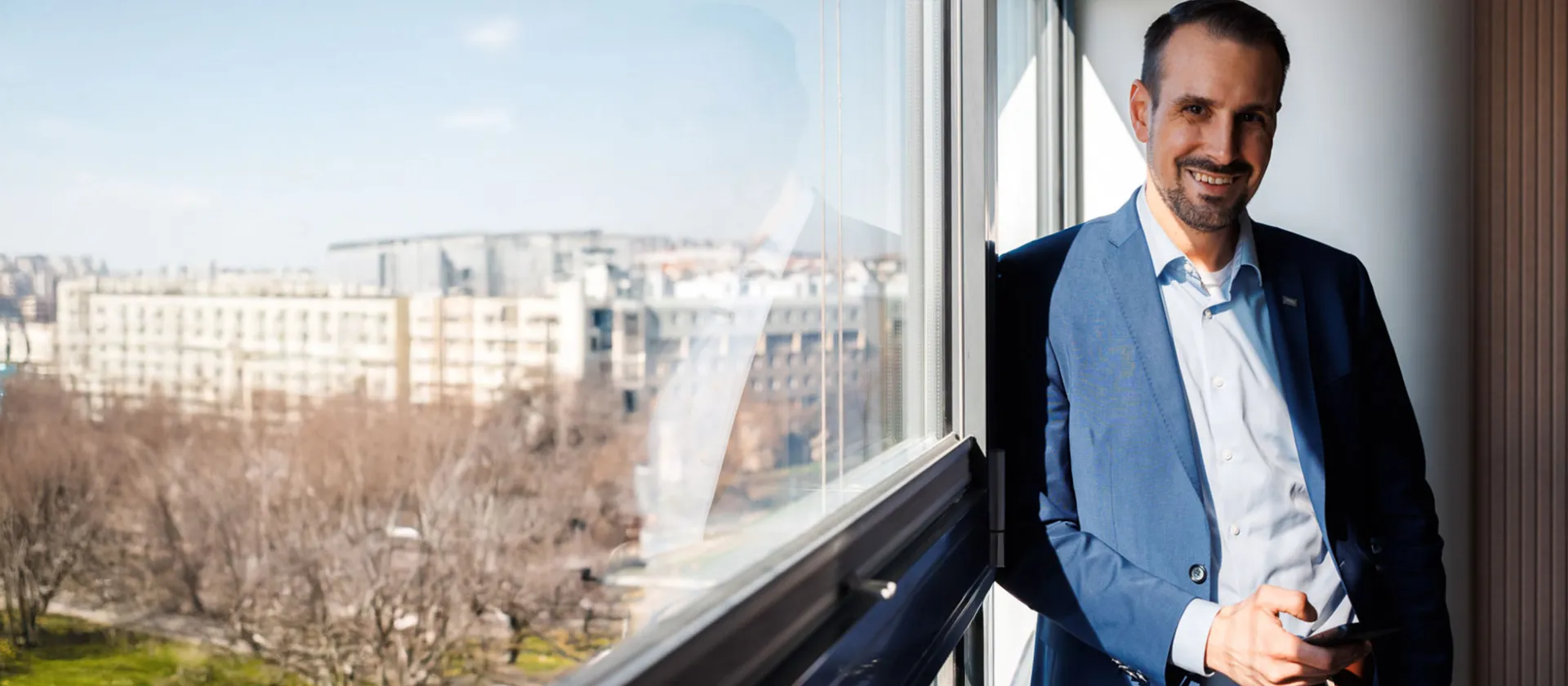 Business professional in a blue suit standing by an office window, holding a smartphone, with a panoramic city view in the background