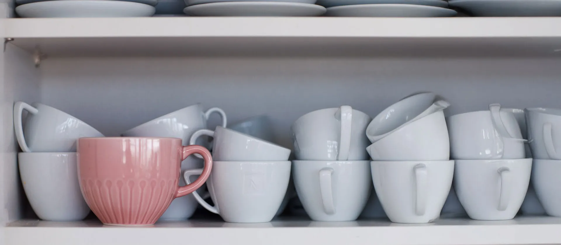  Close-up of white coffee cups stacked on a shelf with one standout pink cup, symbolizing individuality and innovation.