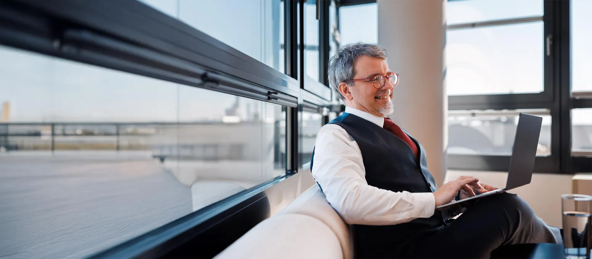 Confident senior executive working on a laptop in a modern office lounge with large windows, showcasing flexible work culture and leadership approachability.