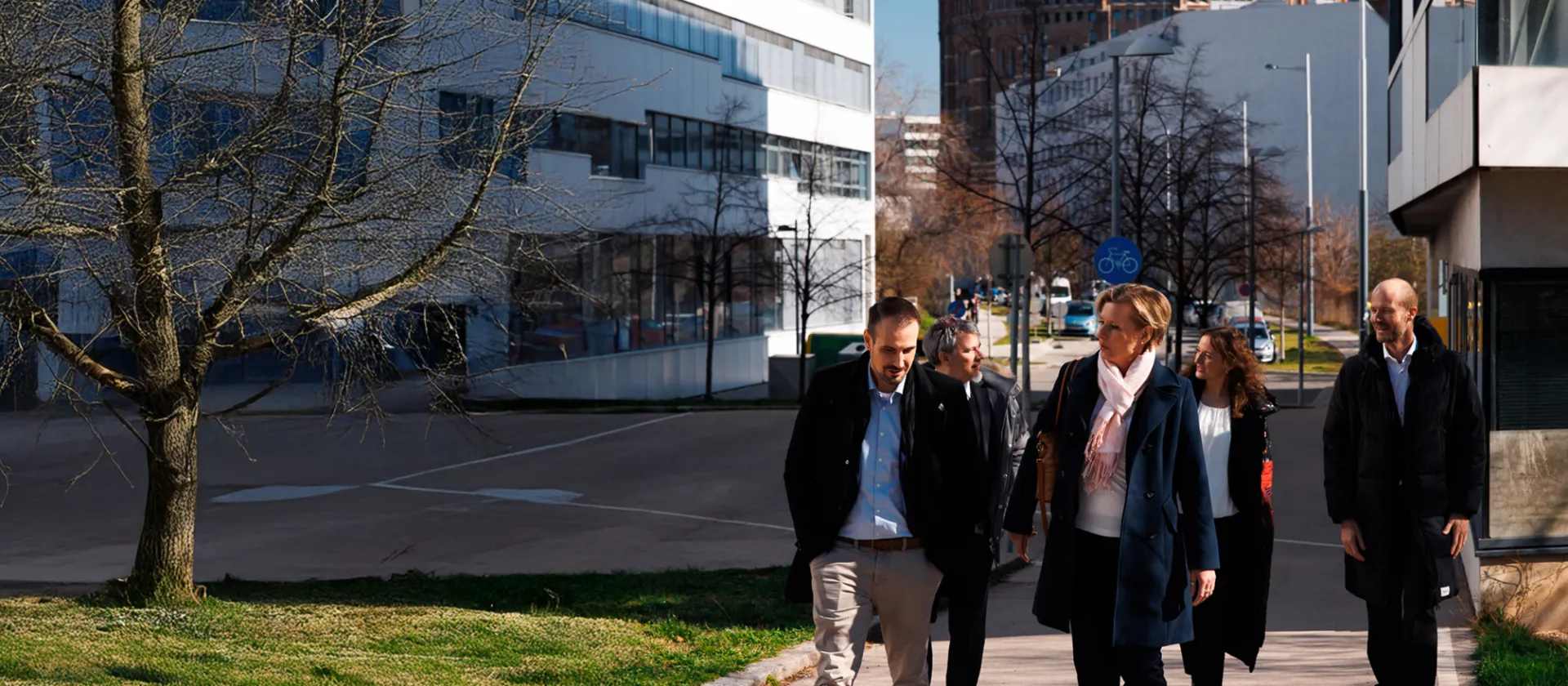 Business professionals walking through a modern urban office district in Vienna, Austria, with the historic Gasometer in the background, on a sunny winter day.