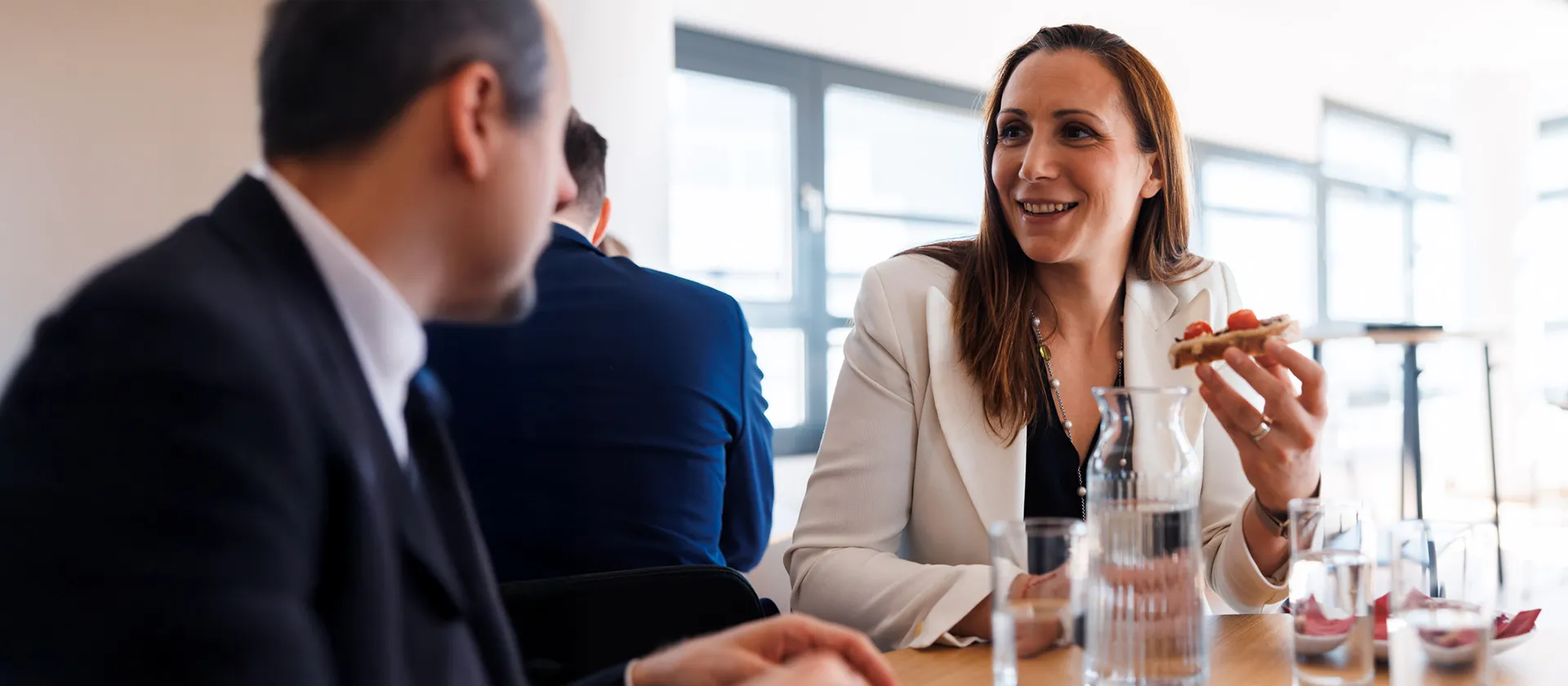 Smiling businesswoman in a white blazer engaging in conversation during an informal company lunch meeting, highlighting a positive and collaborative workplace atmosphere.