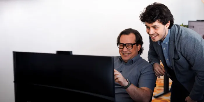 Two colleagues collaborating in front of a computer screen, smiling and discussing project details in a modern office environment.