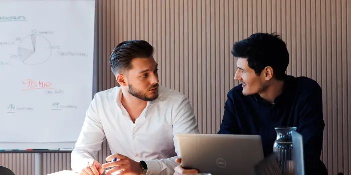 Two young professionals discussing a business strategy in a meeting room, with a flip chart showing a market analysis in the background.
