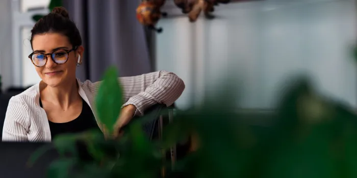 woman with glasses working remotely, smiling while engaged in a virtual meeting, surrounded by greenery in a modern office setting.