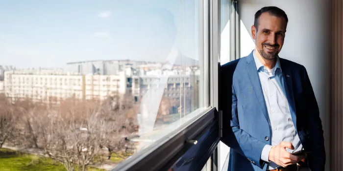 Business professional in a blue suit standing by an office window, holding a smartphone, with a panoramic city view in the background