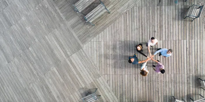 Aerial view of a diverse business team standing in a circle on a wooden deck, joining hands in the center to symbolize collaboration, innovation, and shared goals.