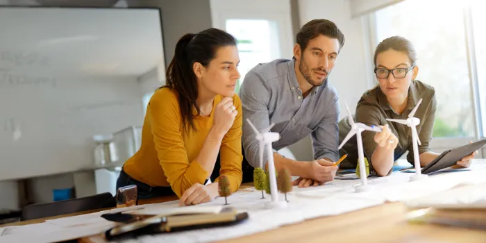 Three professionals collaborating on a wind energy project, analyzing miniature wind turbines and sustainability concepts in a bright, modern office environment.