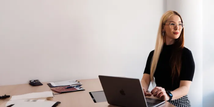 Young professional woman with long hair working on a laptop in a bright, modern office, surrounded by documents and creative materials.