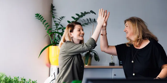 Two smiling female colleagues give each other a high-five in a bright modern office, celebrating a successful collaboration in a positive work environment.