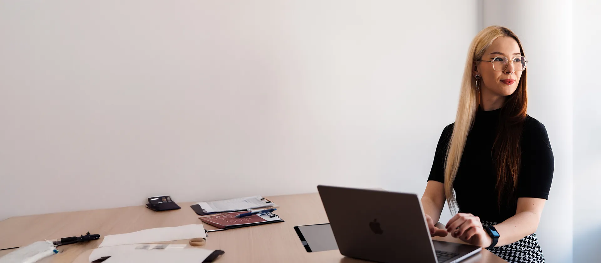 Young professional woman with long hair working on a laptop in a bright, modern office, surrounded by documents and creative materials.