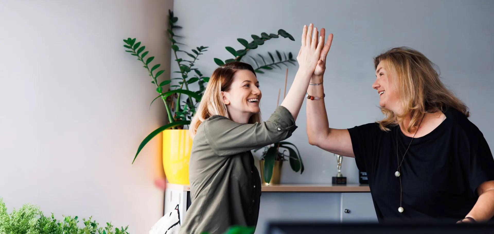 Two smiling female colleagues give each other a high-five in a bright modern office, celebrating a successful collaboration in a positive work environment.