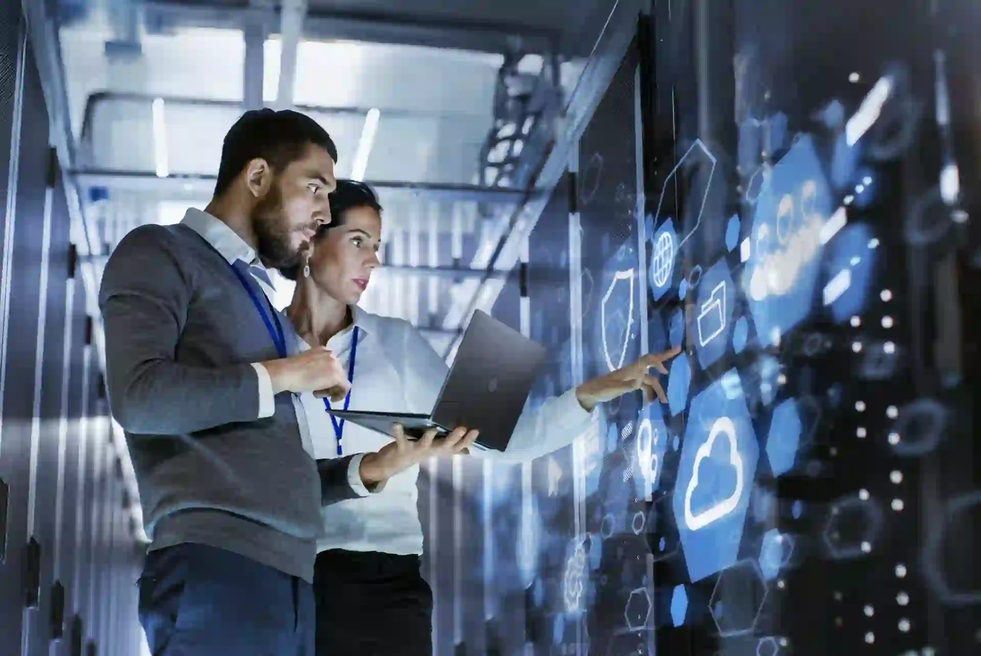 Zwei IT-Fachkräfte in einem Serverraum analysieren Daten auf einem holographischen Display mit Cloud- und Sicherheitssymbolen.